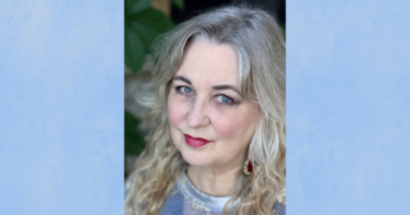Close-up portrait of a smiling woman with wavy blonde hair, blue eyes, and red lipstick wearing a red-eared earring against a soft blue background.